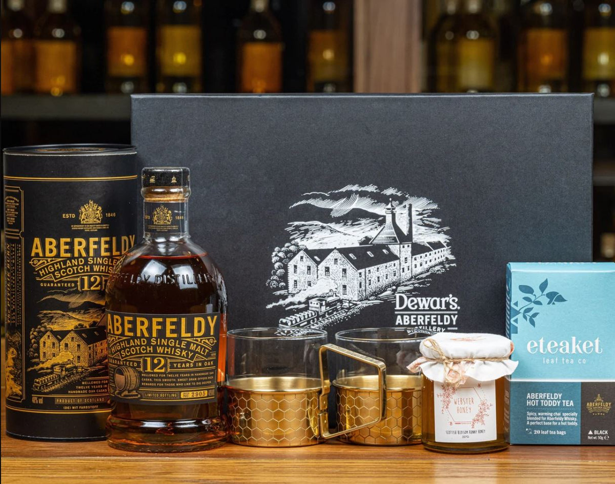 A curated gift set on a wooden surface featuring a bottle of Aberfeldy 12 Year Single Malt Scotch whisky, a matching whisky cylinder, a black branded box, two gold-toned tumblers, a small jar candle, and a blue box of bespoke eteaket Aberfeldy Hot Toddy Tea, with blurred whisky bottles in the background.