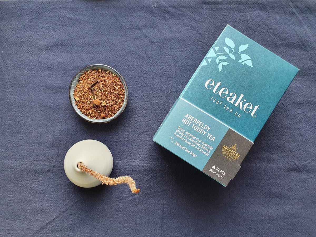 A teal and white box of eteaket x Aberfeldy custom blend: Aberfeldy Hot Toddy Tea displayed on a textured navy blue fabric background, alongside a small bowl of loose leaf tea blend.