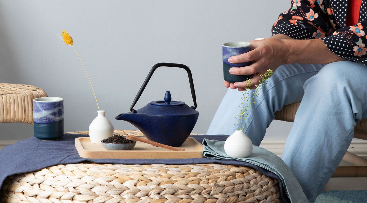 A lifestyle scene showing a person sitting and holding a dark blue ceramic cup, with a matching dark cast iron teapot and other tea accessories arranged on a woven round table. Representing eteaket's tea events and experiences - shared cuppas for team building & corporate wellness. 