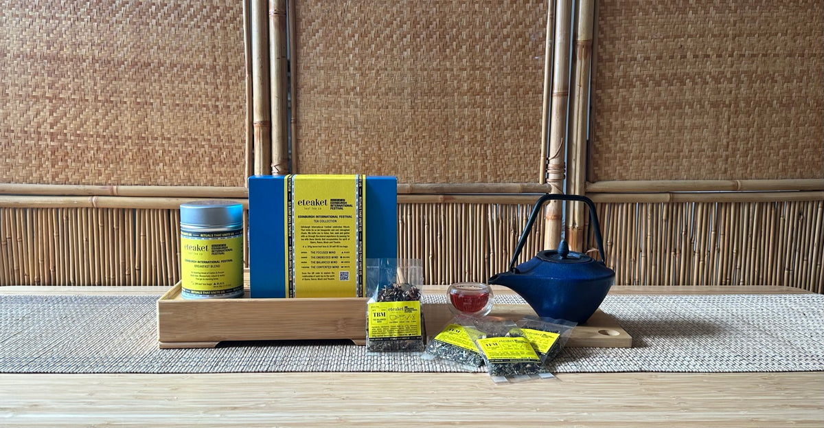 A bespoke tea range crated for Edinburgh International Festival, 'rituals that unite us' A tea set arrangement on a woven mat, featuring a blue teapot, a jar with a yellow branded label, a blue and yellow box, loose tea bags, and small yellow sachets on a bamboo table against a woven bamboo wall.
